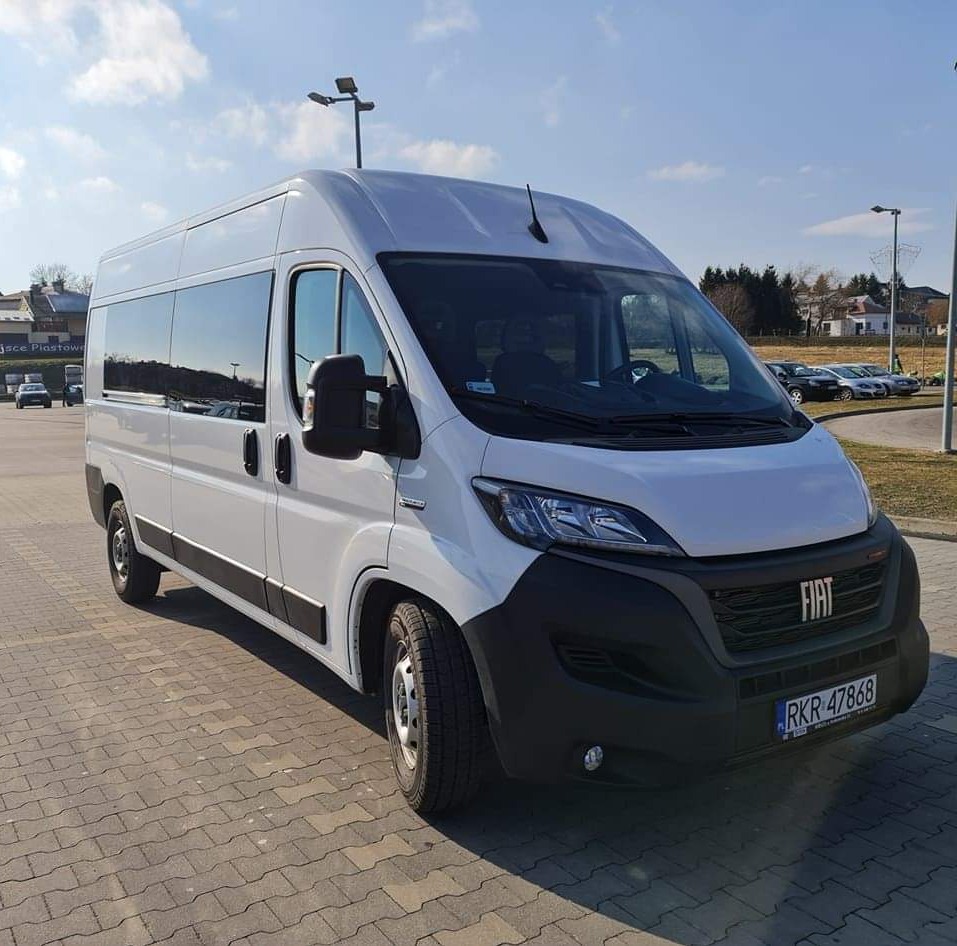 Biały, wysoki bus Fiat Ducato z przyciemnianymi szybami, zaparkowany na kostce brukowej w słoneczny dzień.