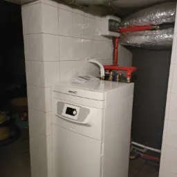 Biały piec gazowy marki Brötje Heizung w kotłowni z instalacją hydrauliczną, widoczne rury w kolorze czerwonym i srebrnym, płytki na ścianach.