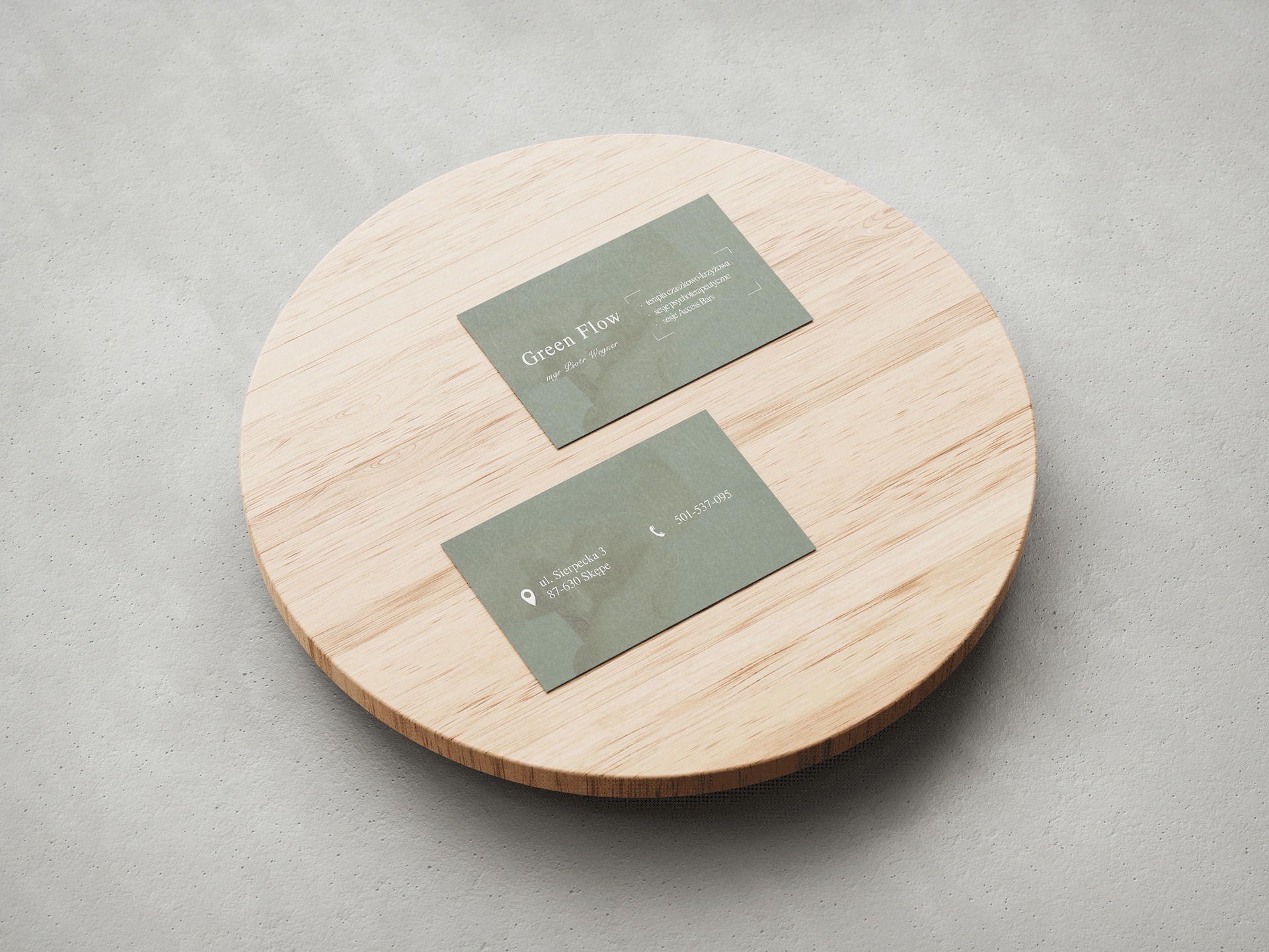 Dwie wizytówki w odcieniach zieleni z danymi kontaktowymi firmy Green Flow, ułożone na okrągłej, drewnianej podstawce na tle szarej, teksturowanej powierzchni.