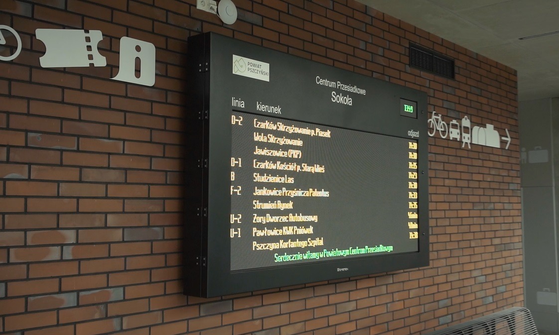 Wyświetlacz LED z dynamiczną informacją pasażerską na ceglanej ścianie w centrum przesiadkowym, prezentujący rozkład jazdy autobusów i linii, obok piktogramy komunikacji miejskiej.