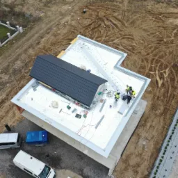 Widok z góry na ekipę w jaskrawych kamizelkach montującą dach z czarnej dachówki na budynku z płaskim dachem pokrytym srebrną folią, obok zaparkowane busy i teren budowy.