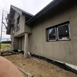 Surowa elewacja nowo budowanego domu jednorodzinnego z widocznymi rusztowaniami, oknami z folią ochronną i częściowo wykonanym drenażem przy fundamencie.