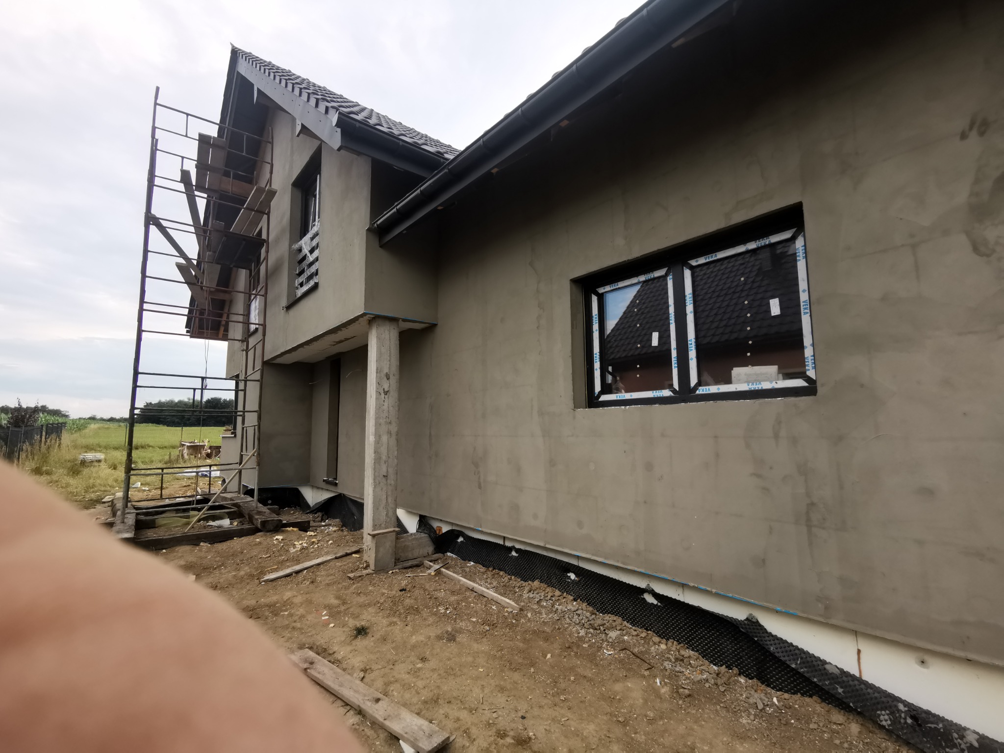 Surowa elewacja nowo budowanego domu jednorodzinnego z widocznymi rusztowaniami, oknami z folią ochronną i częściowo wykonanym drenażem przy fundamencie.