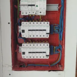 Rozdzielnica elektryczna z zabezpieczeniami różnicowoprądowymi i nadprądowymi Schneider, zamontowana w czerwonej obudowie w ścianie z widocznymi śladami po instalacji.