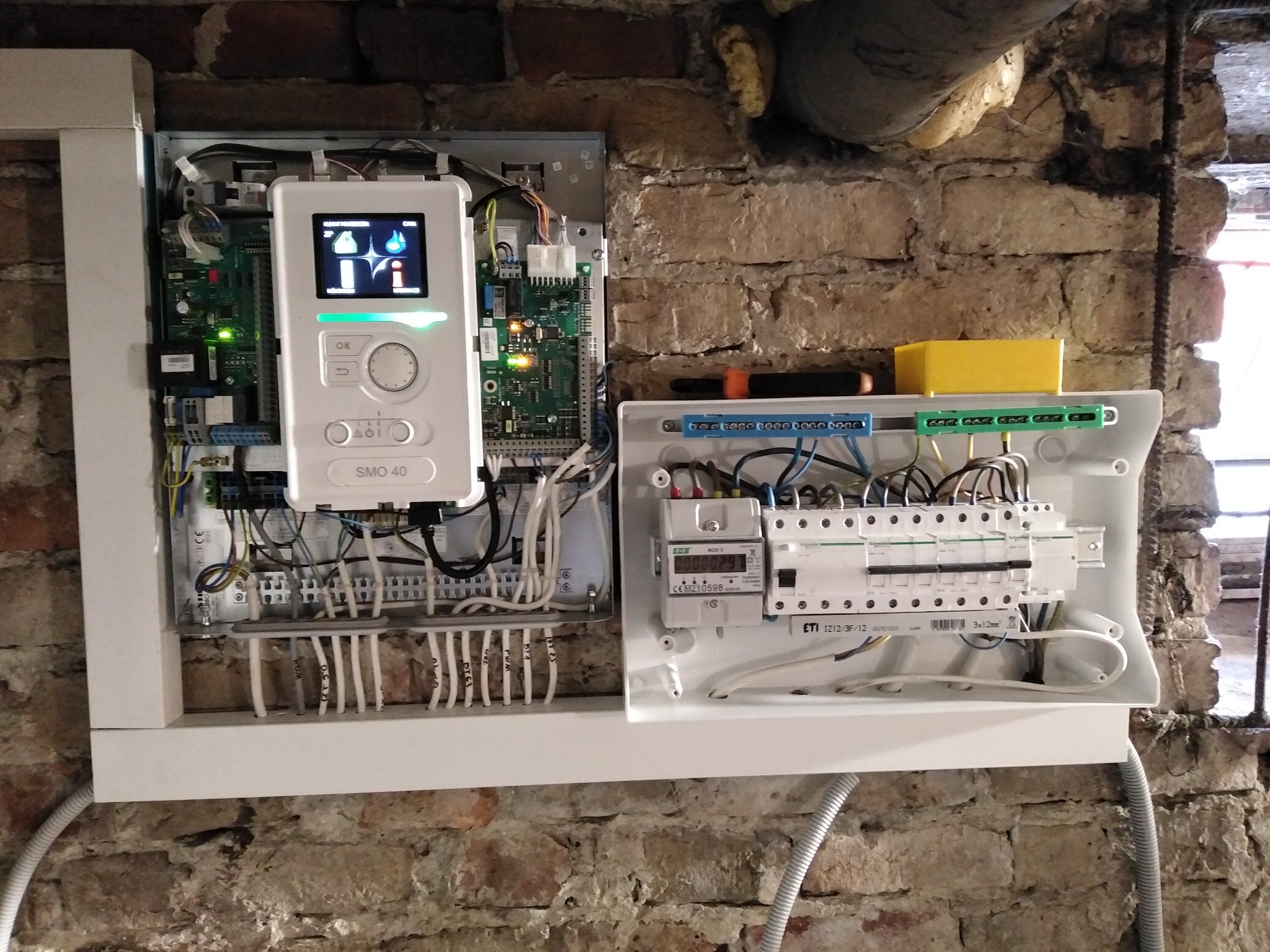 Rozdzielnica elektryczna z panelem sterowania SMO-40 zamontowana na ceglanej ścianie, widoczne przewody, bezpieczniki i licznik energii.