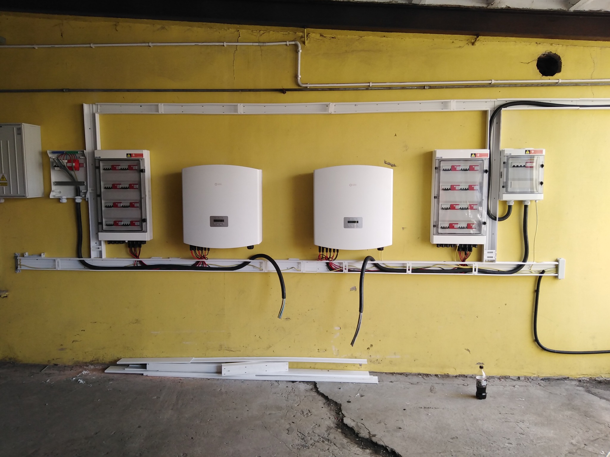 Żółta ściana garażu z zamontowanymi dwoma białymi falownikami solarnymi, rozdzielnicami elektrycznymi i przewodami w korytkach kablowych. Widoczna butelka po napoju na podłodze.