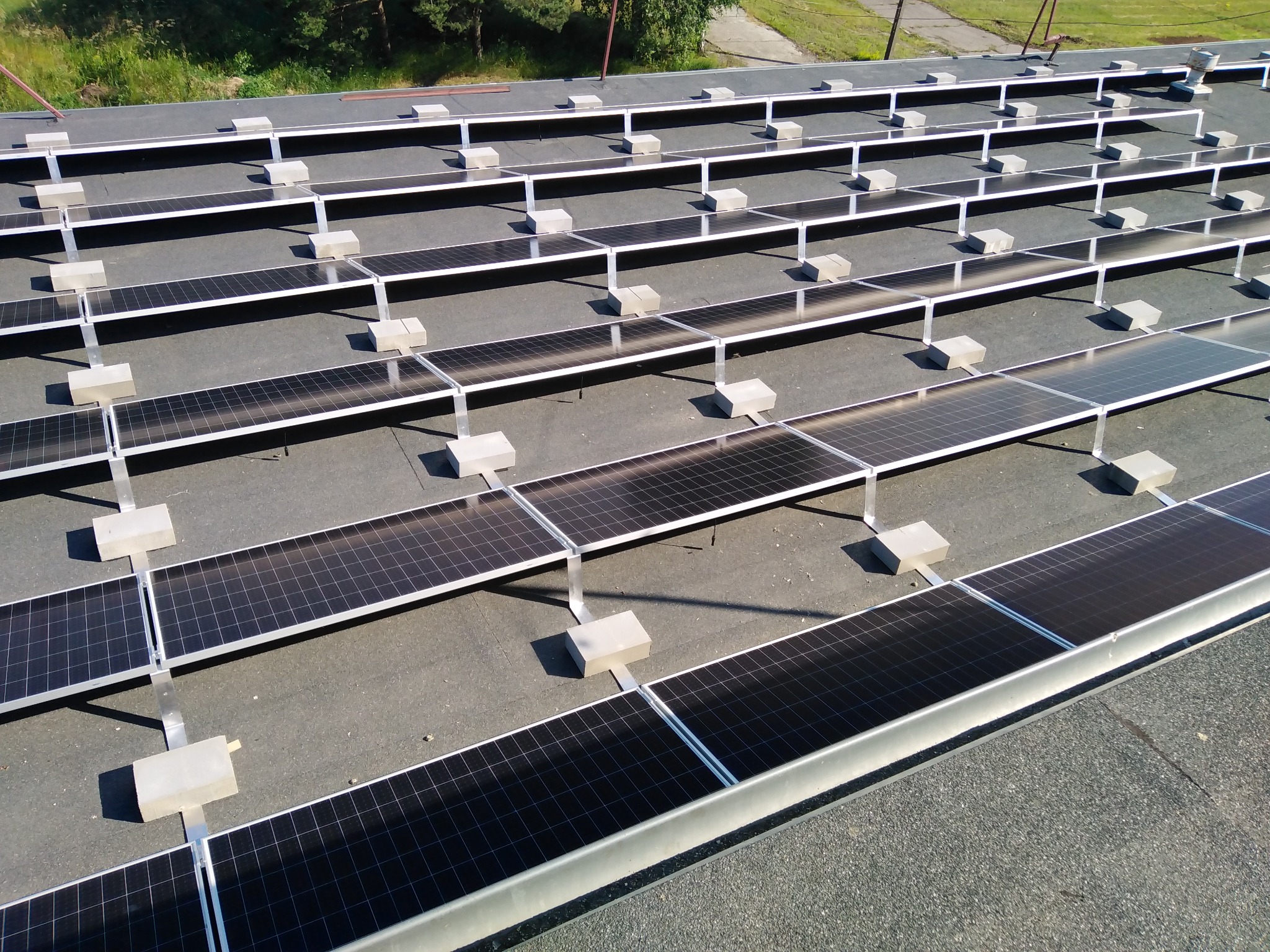 Widok z góry na rząd paneli słonecznych zamontowanych na płaskim dachu z szarym pokryciem, panele ustawione w rzędach na metalowych konstrukcjach z betonowymi obciążnikami, w tle widoczne drzewa.