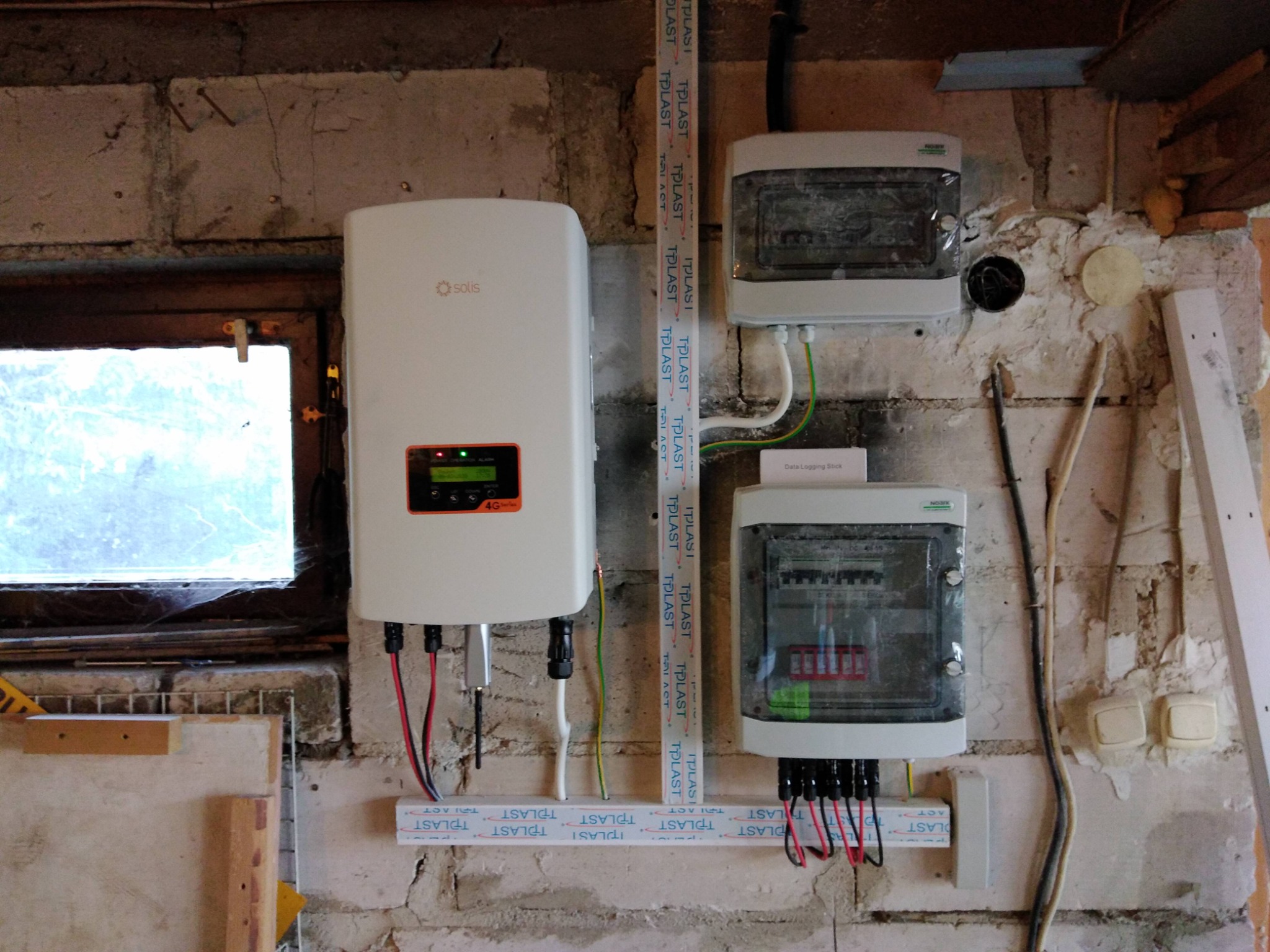 Biały inwerter Solis z podłączonymi przewodami, zamontowany na surowej ścianie z cegieł obok okna, wraz z dwoma skrzynkami rozdzielczymi na ścianie. Widoczne przewody w białych listwach TPLAST.