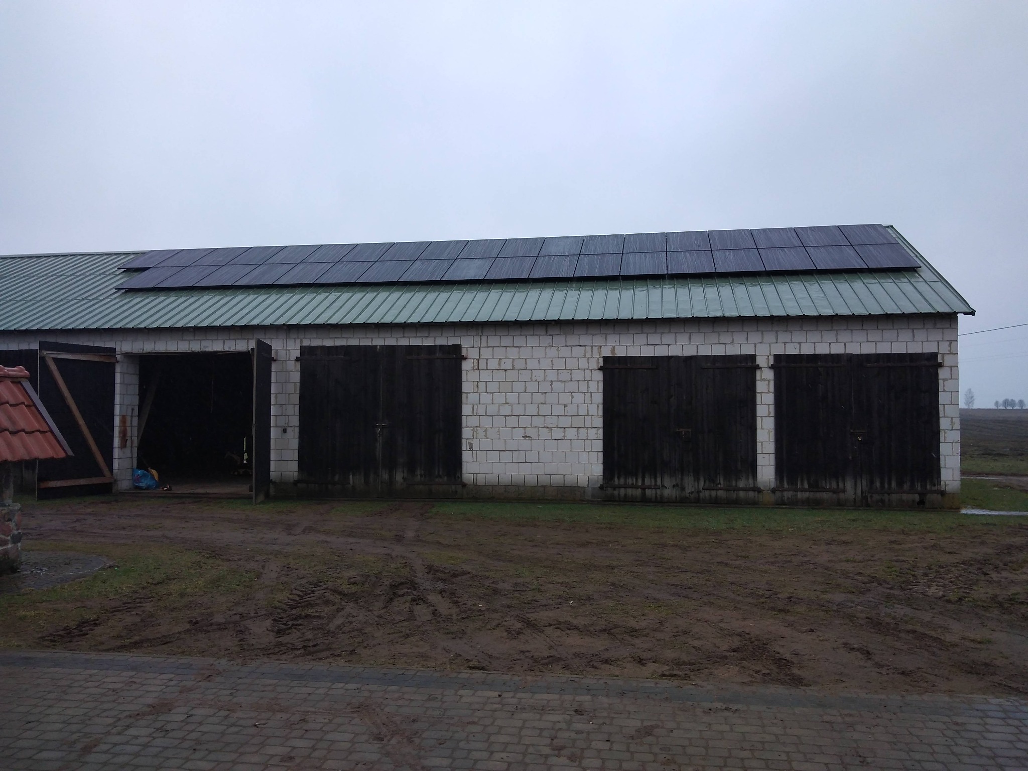Budynek gospodarczy z białej cegły z zamontowanymi panelami fotowoltaicznymi na zielonym, blaszanym dachu, widoczne otwarte wrota garażowe i błotniste podłoże