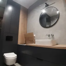 Nowoczesna łazienka z szarą płytką, czarnymi szafkami i blatem z jasnego drewna; widoczna toaleta wisząca i okrągłe lustro z czarną ramą.