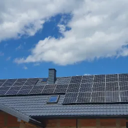 Panele słoneczne na dachu domu w trakcie budowy, widoczne cegły i dachówka w kolorze grafitowym, błękitne niebo z białymi chmurami w tle.