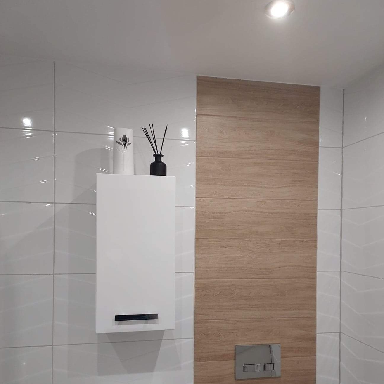 Wykończona łazienka z białymi, błyszczącymi płytkami, drewnopodobną ścianą akcentującą, wiszącą białą szafką oraz dekoracyjnymi dyfuzorami zapachowymi.