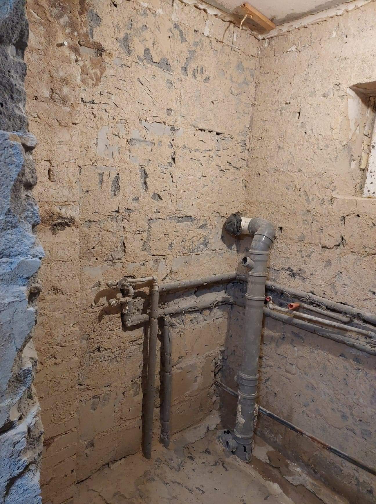 Surowe wnętrze w trakcie prac: widoczne stare, nierówne ściany z odsłoniętą cegłą i resztkami tynku, szare rury instalacji wodnej i kanalizacyjnej, fragment okna w górnym rogu, drewniana belka...