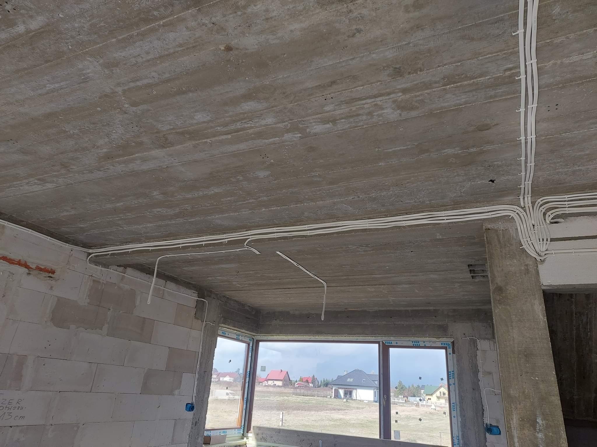 Surowy betonowy strop z widocznymi przewodami elektrycznymi zamocowanymi do sufitu, ściany z bloków betonowych w stanie surowym, okno z widokiem na domy w tle.