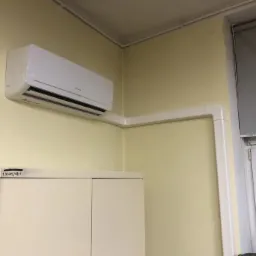 Biały klimatyzator ścienny zamontowany w rogu pomieszczenia biurowego z widocznymi białymi listwami maskującymi przewody, nad szafką z napisem '3'