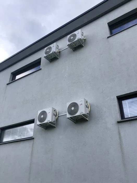 Cztery klimatyzatory zamontowane na szarej ścianie budynku z oknami, widok z dołu na pochmurne niebo.