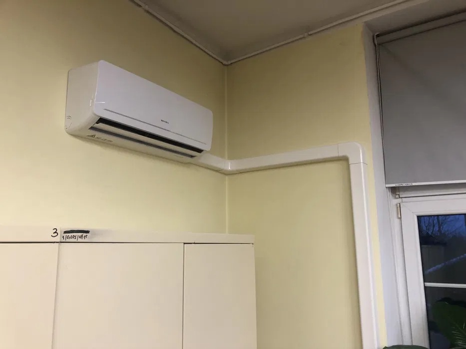 Biały klimatyzator ścienny zamontowany w rogu pomieszczenia biurowego z widocznymi białymi listwami maskującymi przewody, nad szafką z napisem '3'