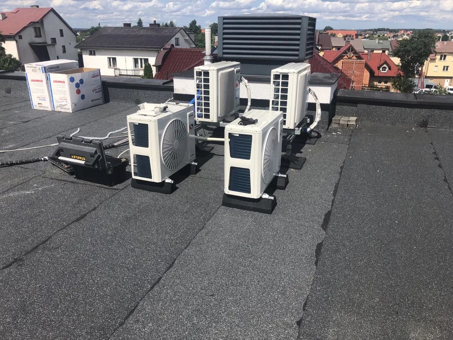 Instalacja kilku klimatyzatorów na dachu budynku, widoczne jednostki zewnętrzne, skrzynka narzędziowa Stanley i kartony w tle.