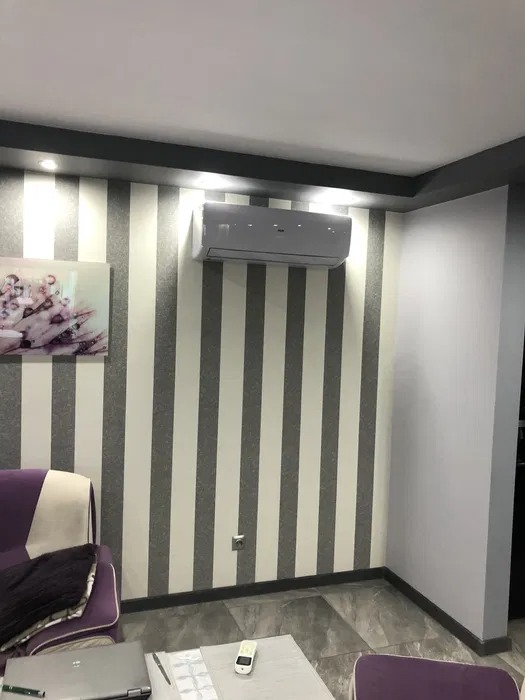 Biały klimatyzator ścienny zamontowany w pokoju z tapetą w szaro-białe paski, widoczny fragment sofy i stolika kawowego.