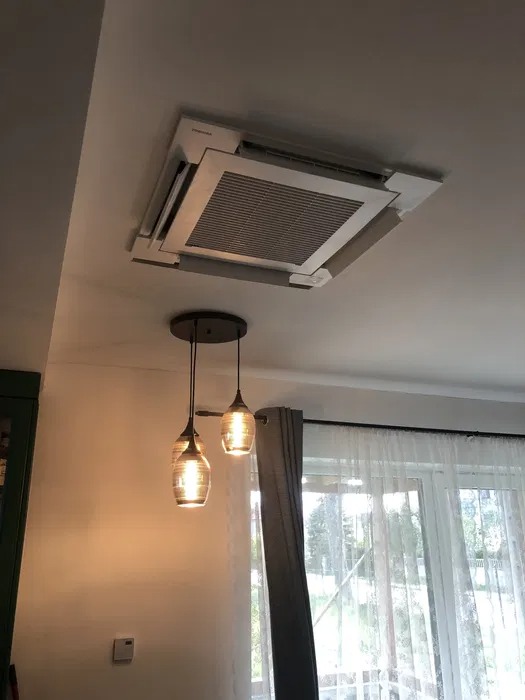 Kasetonowa jednostka klimatyzacyjna marki Toshiba zamontowana w suficie pomieszczenia mieszkalnego, widoczna lampa wisząca z trzema szklanymi kloszami oraz okno z firankami.
