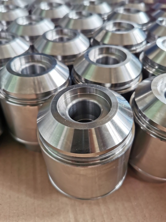 Partie precyzyjnie obrobionych, metalowych elementów cylindrycznych z kołnierzem i centralnym otworem, ułożonych na tekturowym podłożu.