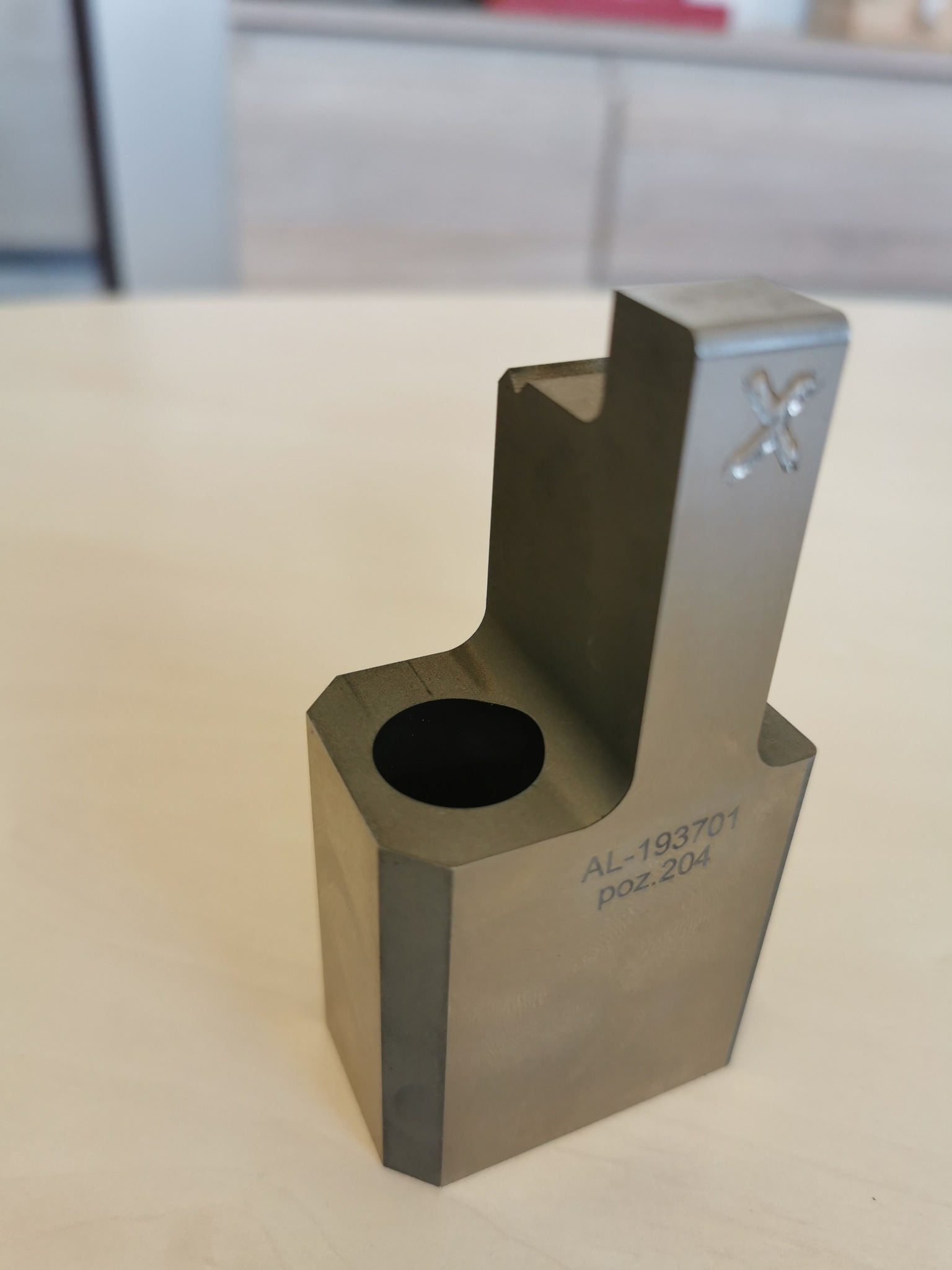 Metalowy element z otworem i wygrawerowanym numerem seryjnym 'AL-193701 poz.204' oraz znakiem graficznym, leżący na jasnym blacie.