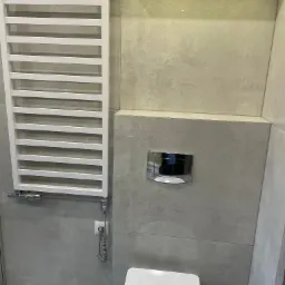 Nowoczesna łazienka z szarymi płytkami, białym grzejnikiem drabinkowym, wiszącą toaletą i podświetleniem LED pod sufitem.