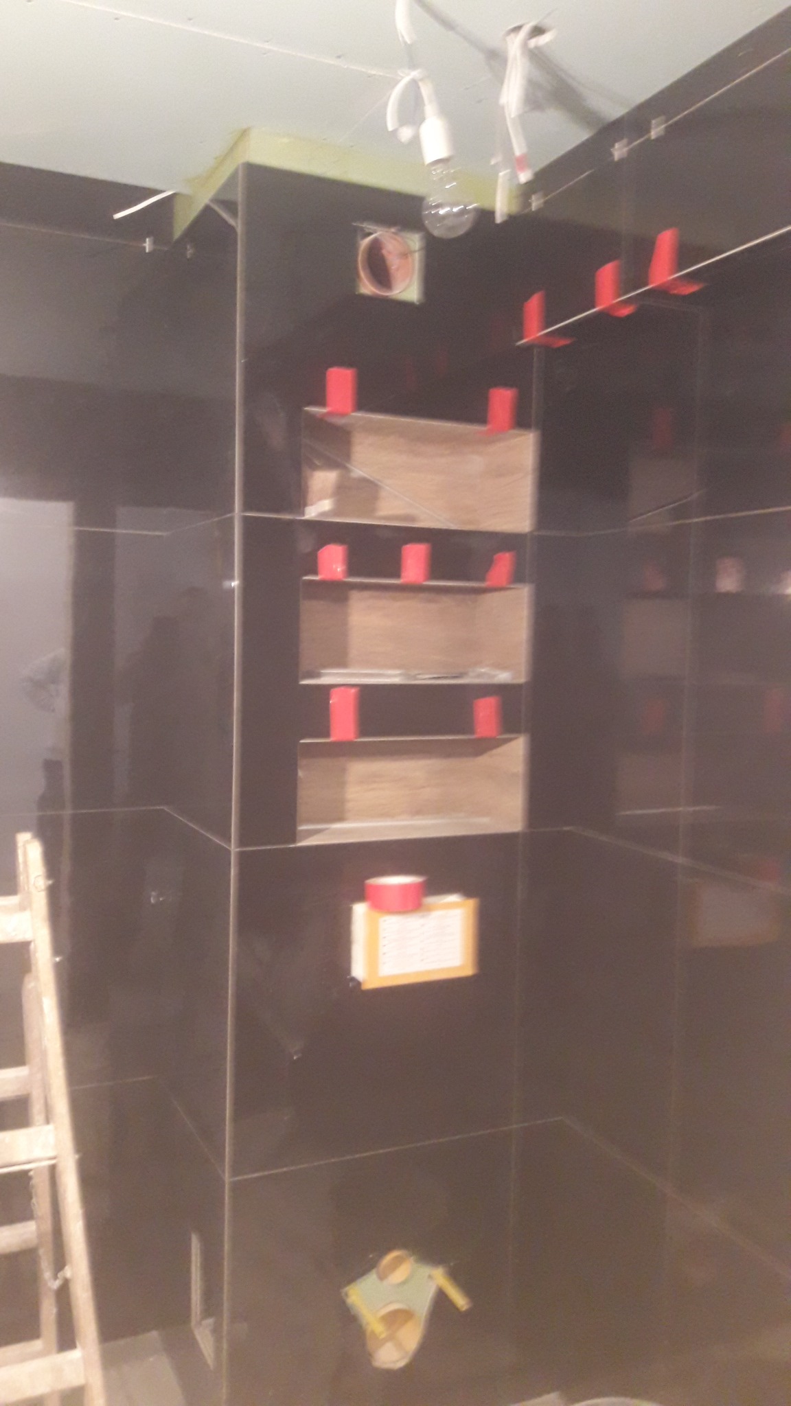 Ciemne, błyszczące płytki ścienne w trakcie montażu, z widocznymi czerwonymi krzyżykami dystansowymi, drewnianymi półkami wnękowymi w trakcie wykańczania i drabinką w rogu pomieszczenia.