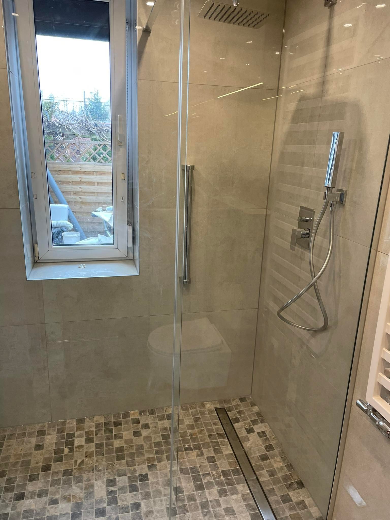 Nowoczesna kabina prysznicowa z odpływem liniowym i oknem, wykończona beżowymi płytkami ściennymi i mozaiką podłogową w odcieniach szarości i brązu, widoczny fragment toalety za szybą.