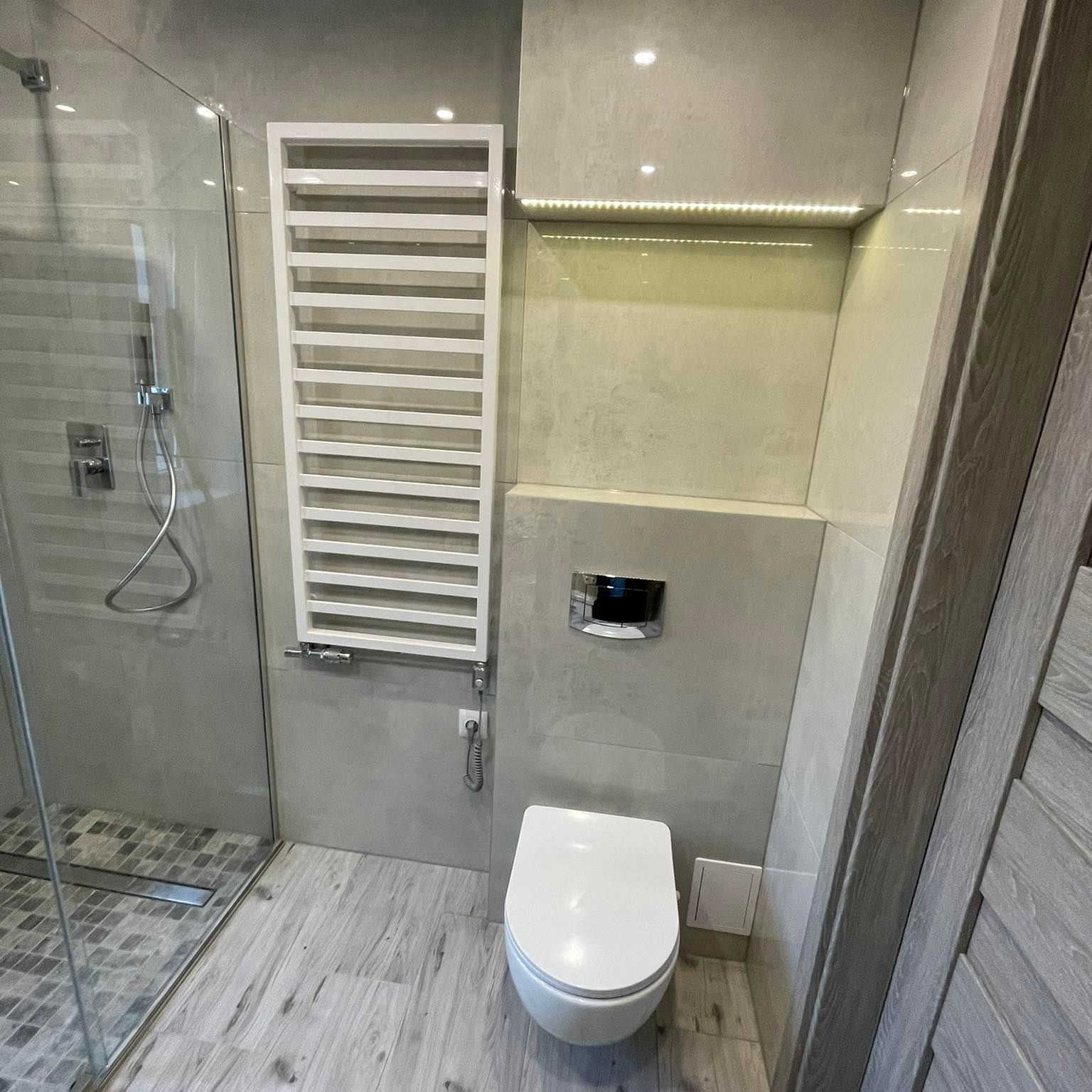 Nowoczesna łazienka z kabiną prysznicową, grzejnikiem drabinkowym i podwieszaną toaletą, wykończona szarymi płytkami imitującymi drewno i kamień, z subtelnym oświetleniem LED.