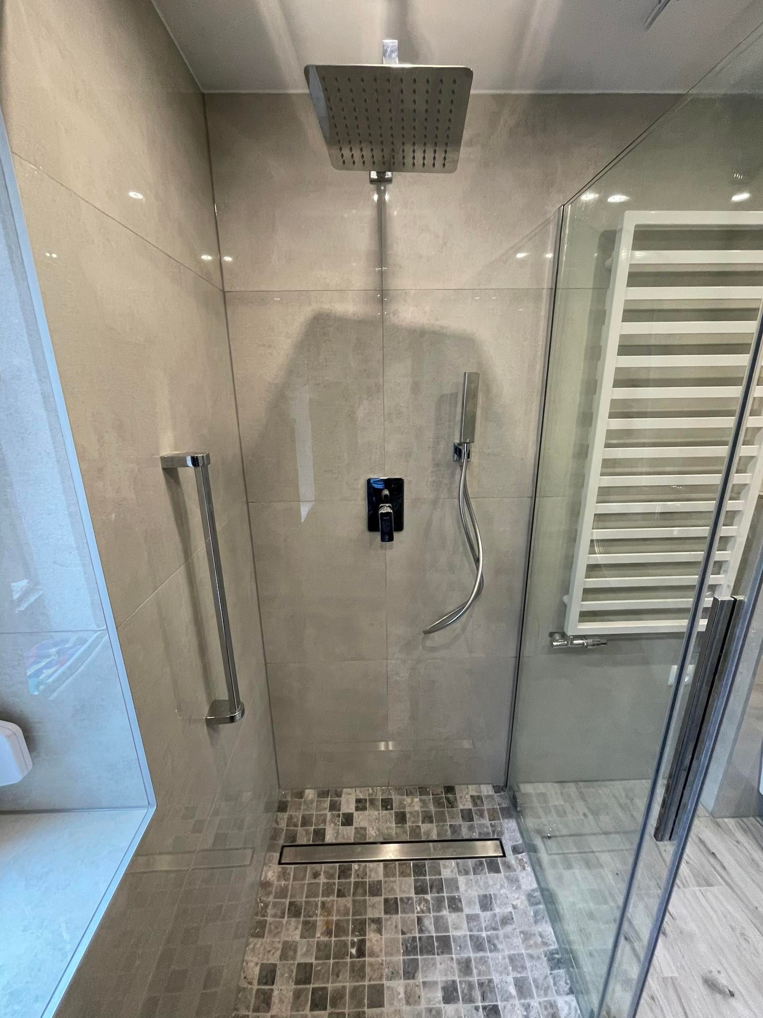 Nowoczesna kabina prysznicowa z deszczownicą, szklaną ścianką i mozaikową posadzką z odpływem liniowym, widoczny grzejnik drabinkowy.