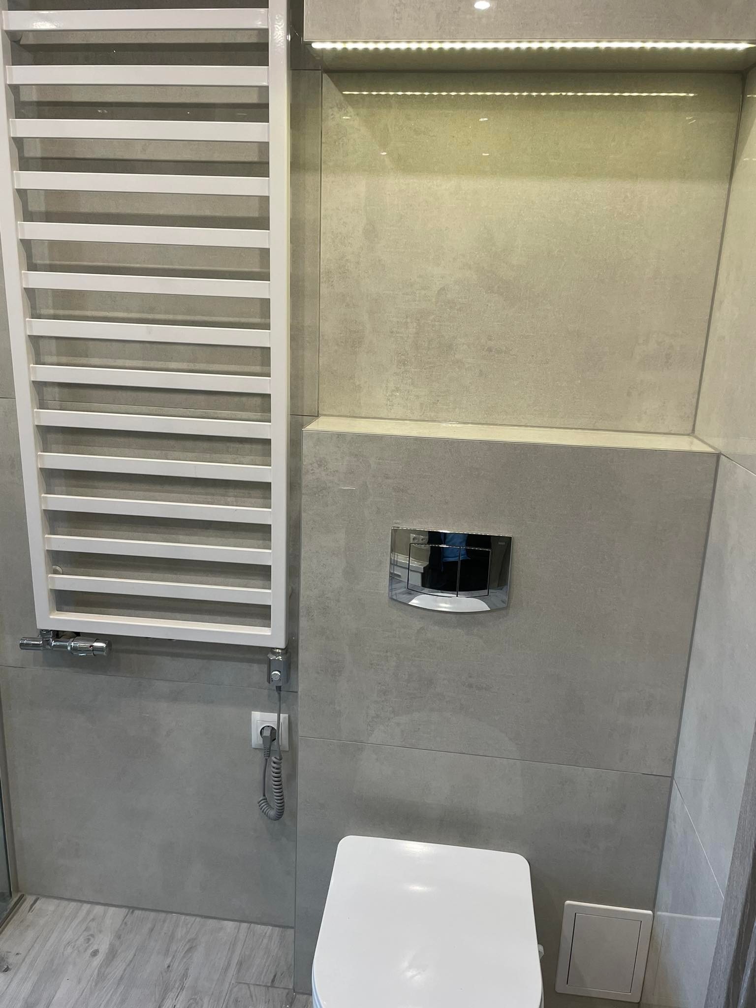 Nowoczesna łazienka z szarymi płytkami, białym grzejnikiem drabinkowym, wiszącą toaletą i podświetleniem LED pod sufitem.