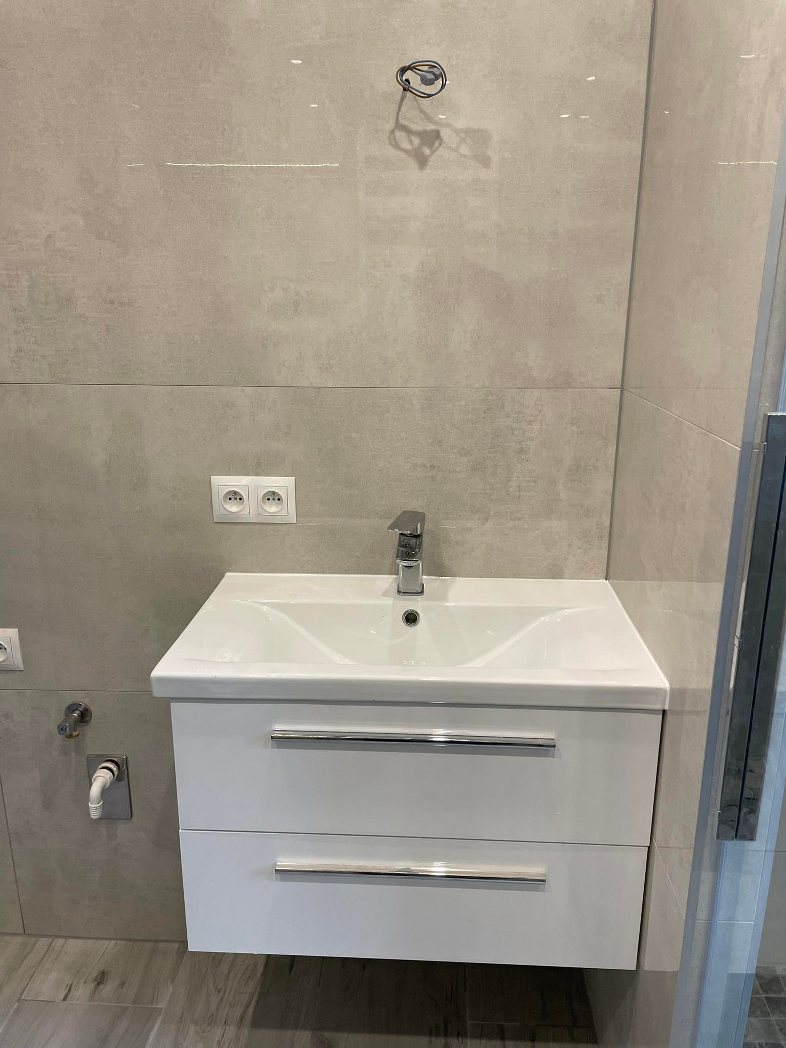 Biała umywalka z szafką i chromowaną baterią na tle szarych, prostokątnych płytek ściennych z widocznymi gniazdkami elektrycznymi i wystającymi przewodami.