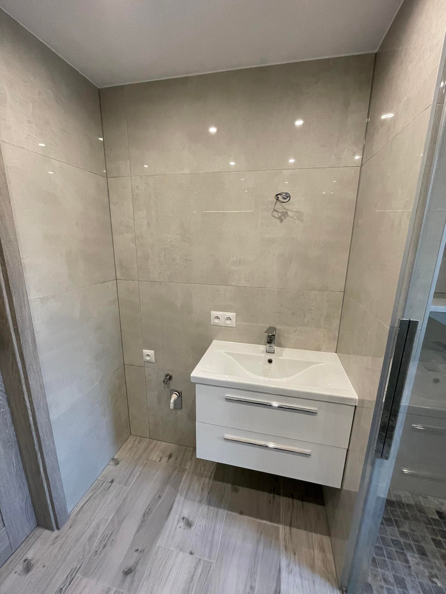 Nowoczesna łazienka z jasnymi, matowymi płytkami imitującymi beton na ścianach, drewnopodobną podłogą, białą szafką pod umywalką z chromowaną armaturą i szklaną kabiną prysznicową.