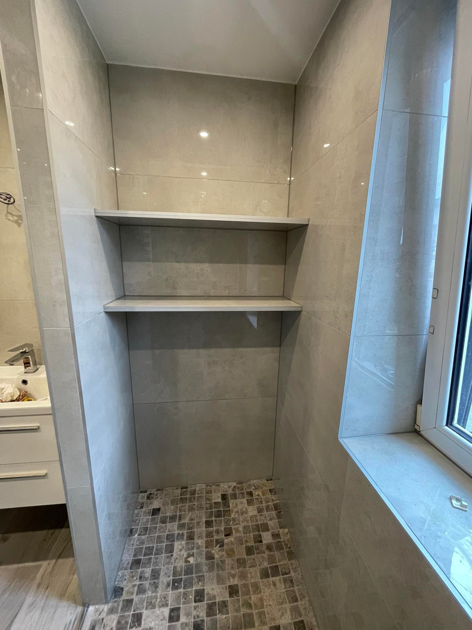 Wykończona wnęka prysznicowa z półkami, wyłożona szarymi płytkami ceramicznymi o nieregularnej fakturze, mozaika podłogowa w odcieniach brązu i szarości, widoczny fragment umywalki z baterią...