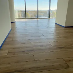 Ułożona podłoga z płytek imitujących drewno w jasnym, nowoczesnym wnętrzu z dużym oknem z widokiem na miasto; ściany zabezpieczone niebieską taśmą malarską.