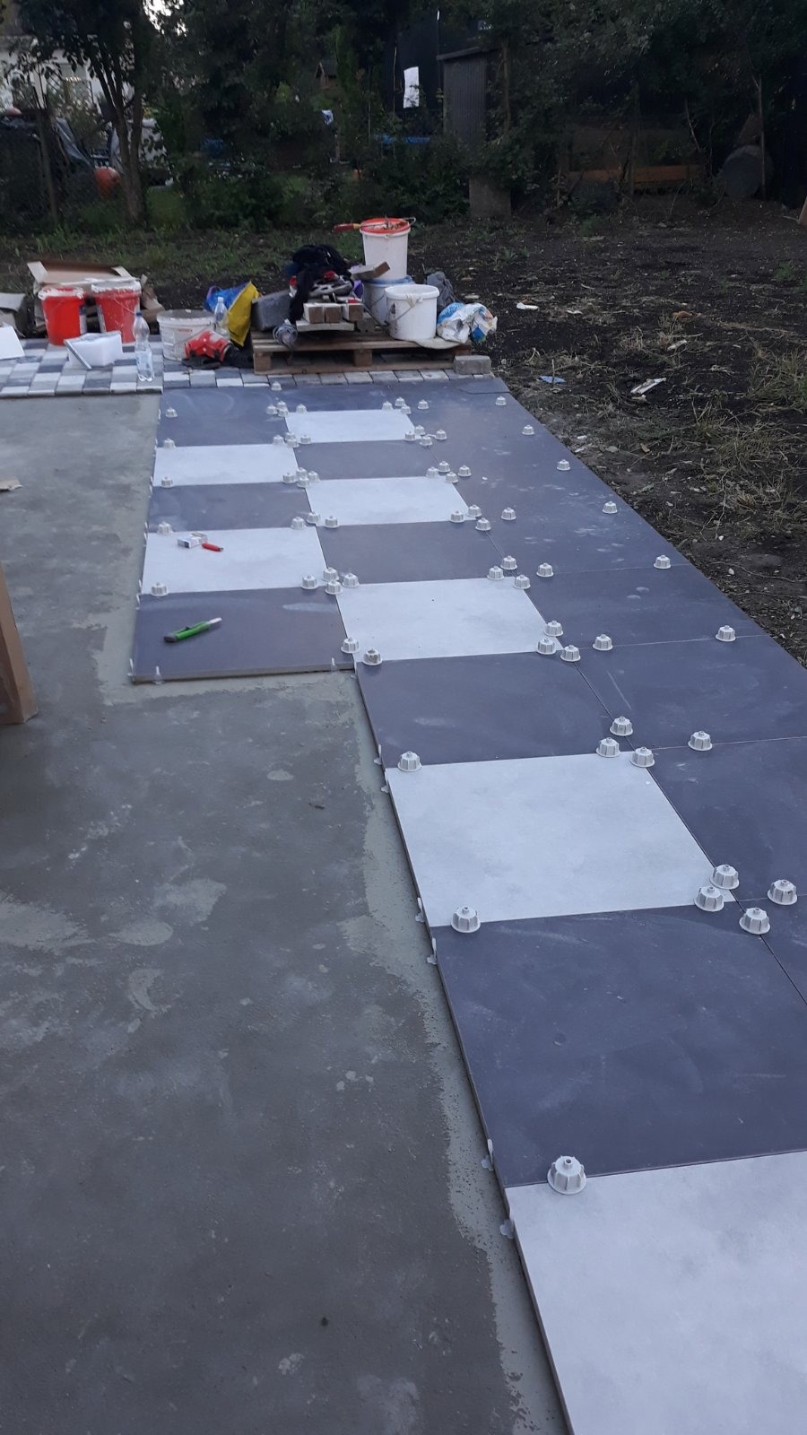 Układanie kwadratowych płytek w szachownicę na zewnątrz, z użyciem systemów poziomowania płytek, widoczny nóż tapicerski i zaplecze budowy.