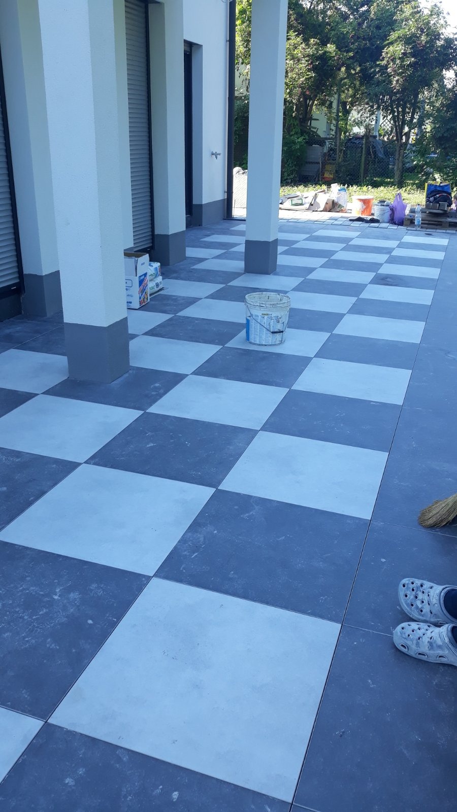 Szachownica z ciemnych i jasnych płytek na tarasie, białe kolumny podtrzymują zadaszenie, wiadro z farbą stoi na środku, a w rogu widoczne klapki crocs.