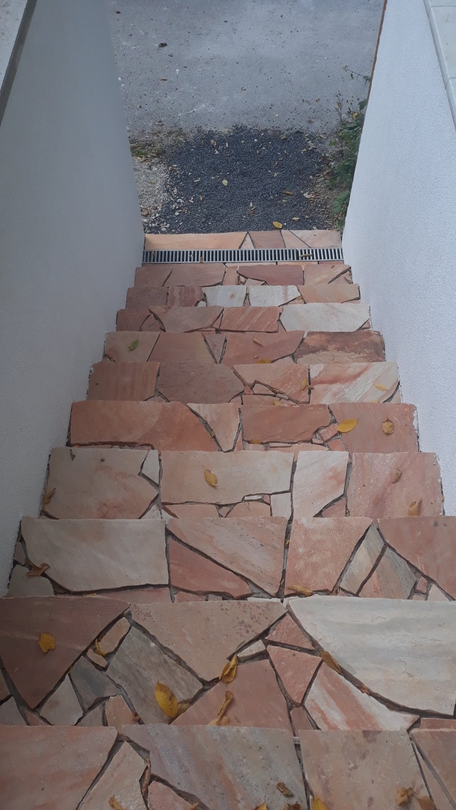 Schody zewnętrzne wyłożone nieregularnymi płytami kamiennymi w odcieniach beżu i różu, z widoczną kratką odpływową na górze i kilkoma opadłymi liśćmi na stopniach.