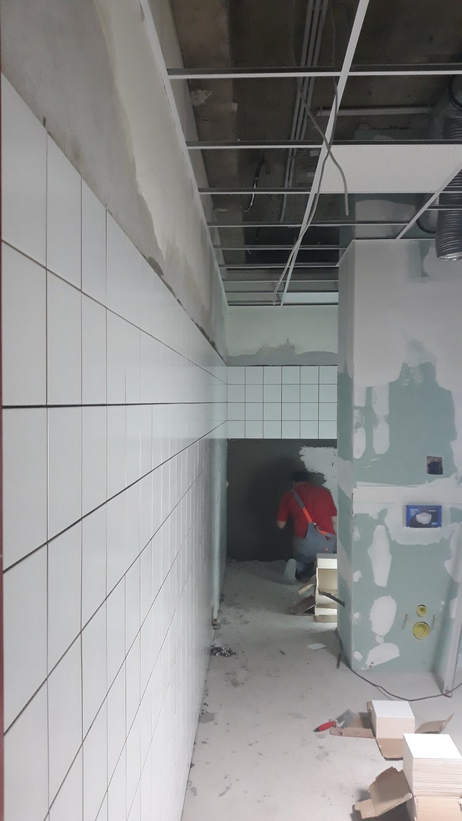 Białe płytki kwadratowe ułożone na ścianie w trakcie remontu łazienki, widoczny fragment sufitu z instalacjami i pracownik w czerwonej koszuli w tle.
