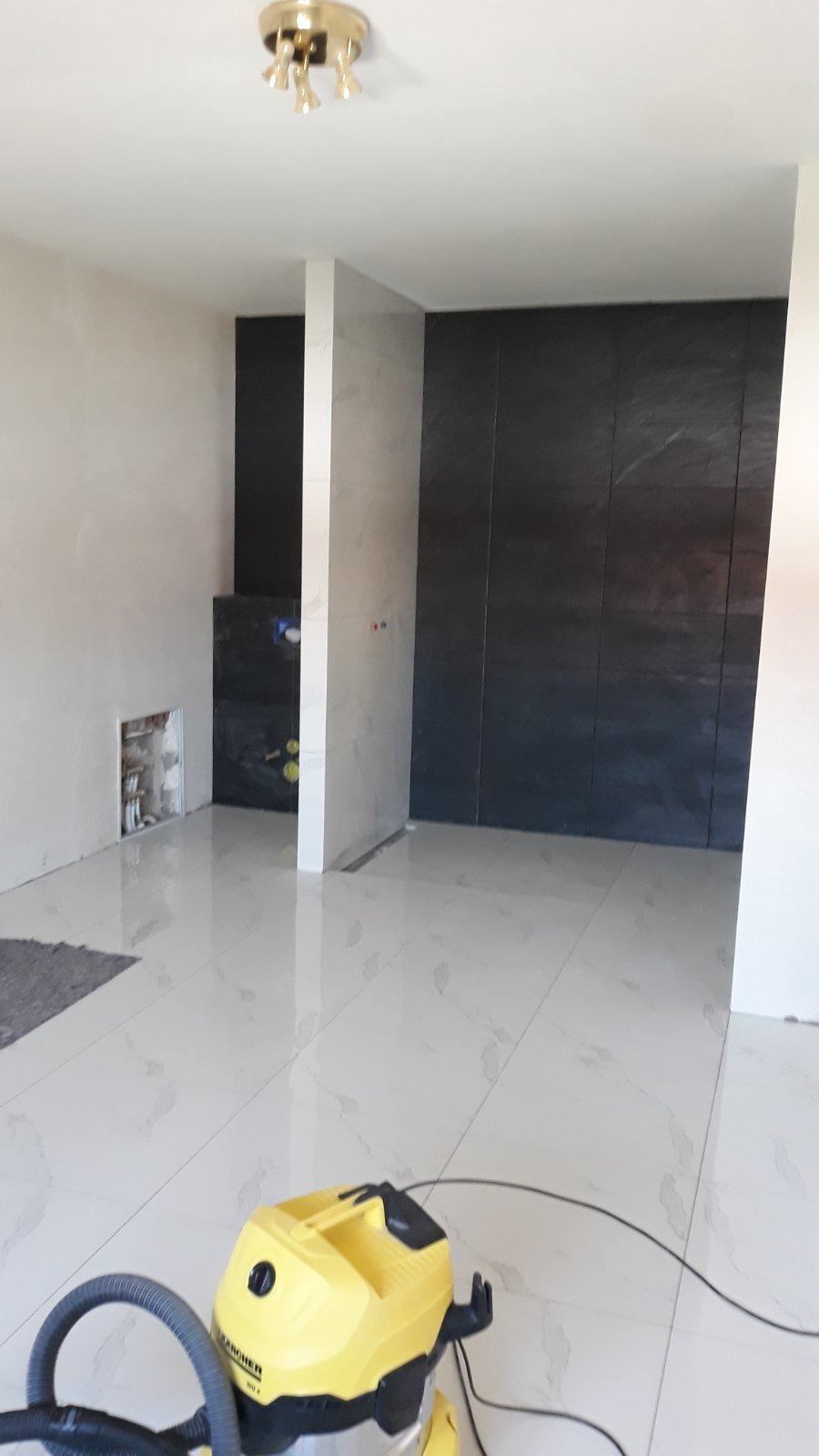 Wnętrze łazienki w trakcie remontu, widoczne czarne płytki na ścianie i białe płytki podłogowe, z żółtym odkurzaczem przemysłowym na pierwszym planie oraz instalacją hydrauliczną w ścianie bez...