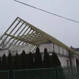 Konstrukcja drewniana dachu w trakcie budowy na domu z białych pustaków, widok zza zielonego ogrodzenia i rzędu iglaków.