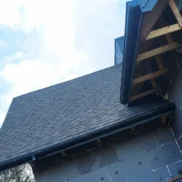 Dach pokryty gontem bitumicznym w kolorze grafitowym, z zamontowanymi rynnami i obróbkami blacharskimi, widoczna izolacja termiczna budynku w trakcie montażu.