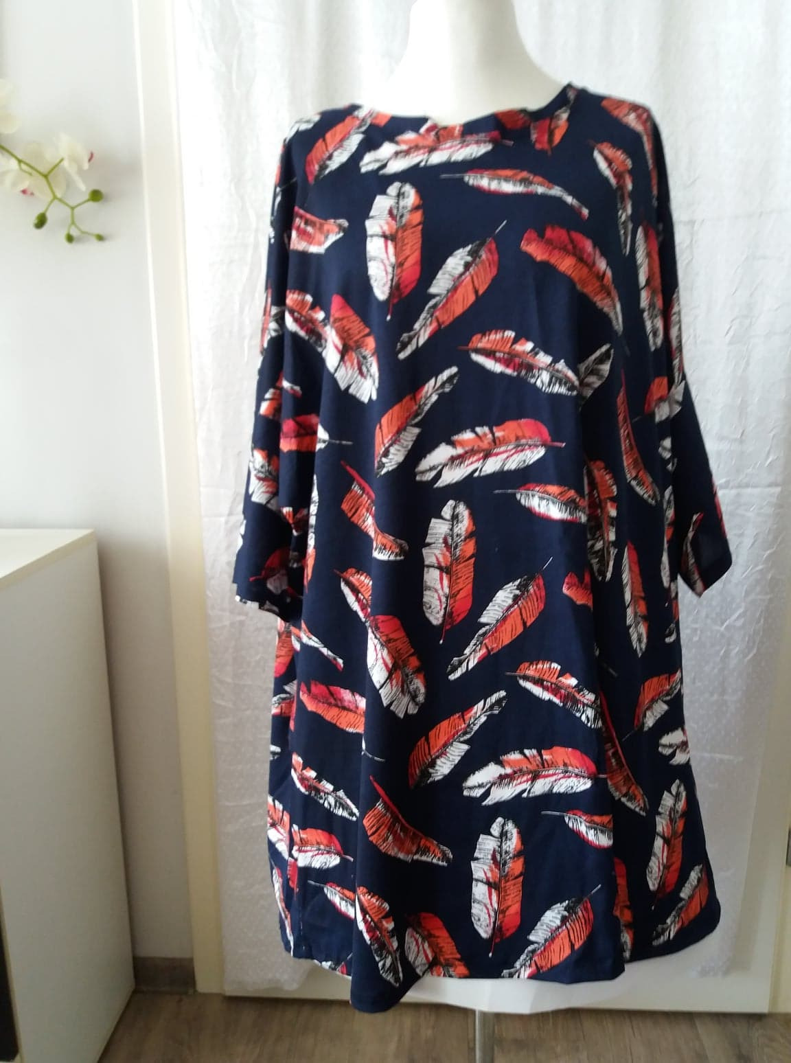 Granatowa sukienka z motywem pomarańczowo-białych piór, prezentowana na manekinie na tle białej ściany i zasłony.