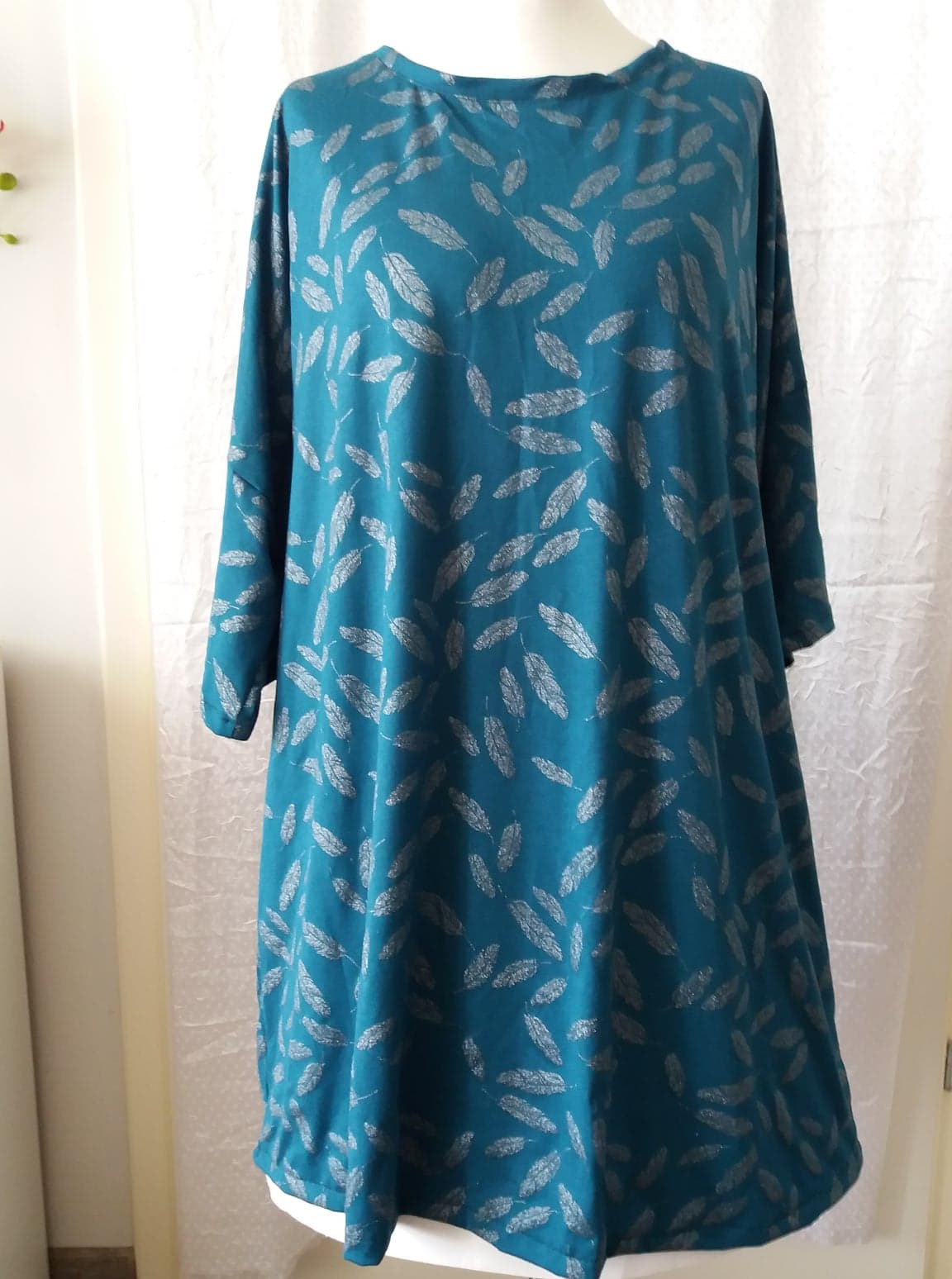 Turkusowa tunika z długim rękawem, pokryta srebrnymi motywami piór, prezentowana na manekinie na tle białej zasłony.