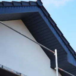 Fragment elewacji budynku z widocznym rusztowaniem, świeżo pomalowana ściana w jasnym kolorze, z ciemnym podbitkiem dachowym i fragmentem dachu na tle błękitnego nieba.