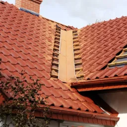 Częściowy demontaż czerwonej dachówki ceramicznej na skośnym dachu domu jednorodzinnego, widoczny komin z cegły, fragment orynnowania i ułożony stos dachówek na rusztowaniu.