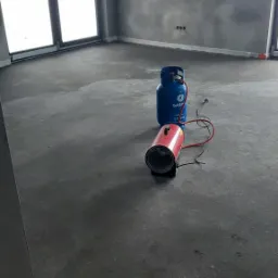 Surowe wnętrze pokoju z świeżo nałożonym tynkiem, widoczny nagrzewnica gazowa i butla gazowa, okna w tle wpuszczają dużo światła dziennego, ściany szare i matowe, z widocznymi puszkami elektrycznymi.