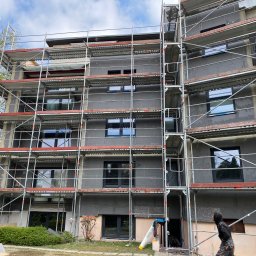 PIERZCHAŁA ELEWACJE BUDYNKÓW - Wielopiętrowy budynek w trakcie ocieplania, z widocznymi rusztowaniami i warstwą izolacyjną na elewacji. Pracownik w stroju roboczym na pierwszym planie.