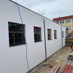 PIERZCHAŁA ROBERT - Biała elewacja budynku z ciemnymi oknami, widoczne rury spustowe i niebieski przewód. W tle budynek z żółtą elewacją i rusztowania. Teren wokół budynku w trakcie prac.